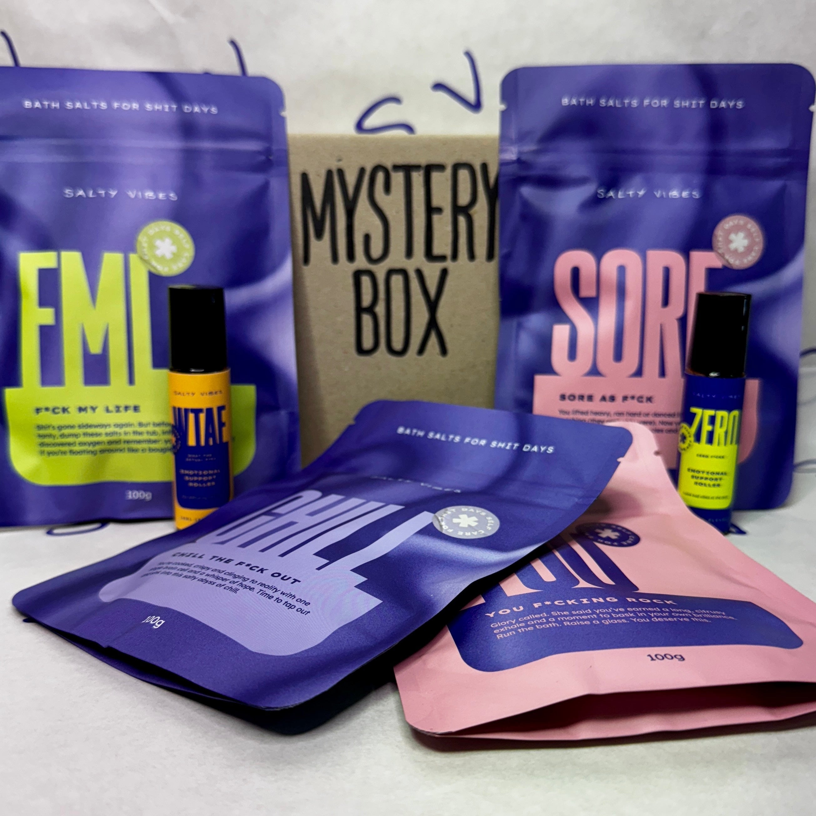 4 bath salts and 2 oil roller bottles in front of a box with Mystery Box written on it.